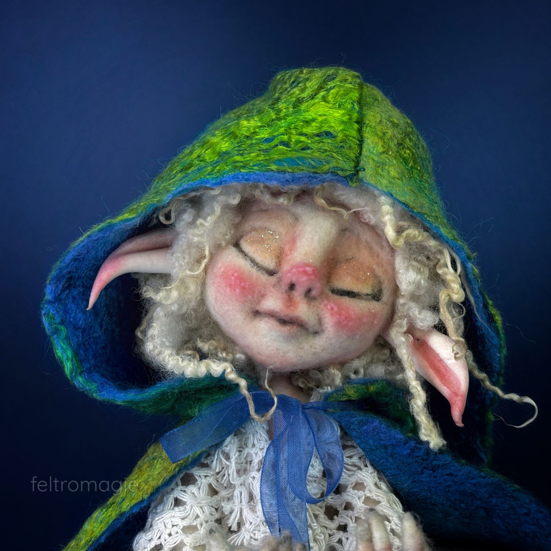 Volto in primo piano della art doll di lana, un'elfa dal volto bianco ceruleo con occhi socchiusi con un grande cappuccio di seta che le avvolge la testa mostrando riccioli bianchi